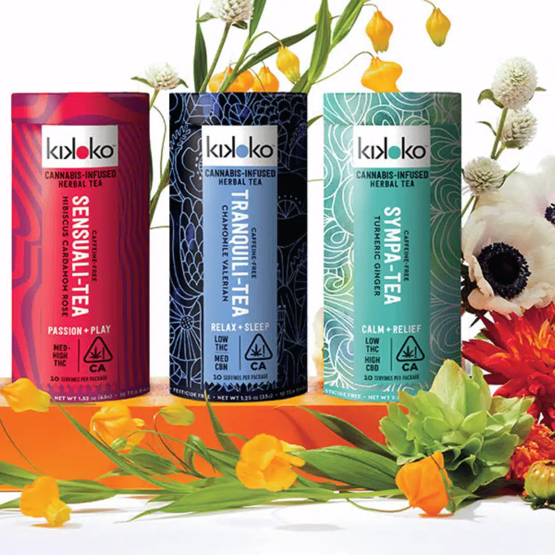 Three Kikoko cannabis-infused herbal tea cans: Sensuali-Tea in red, Tranquili-Tea in blue, and Sympa-Tea in green, elegantly placed among colorful flowers. A bright and refreshing representation of wellness and relaxation.