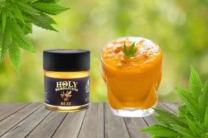 A jar of Holy Herbajuana Buzz cannabis-infused honey sits on a wooden surface next to a vibrant orange smoothie in a glass, garnished with a small cannabis leaf. Fresh cannabis leaves frame the image, with a blurred green background creating a natural and refreshing atmosphere.